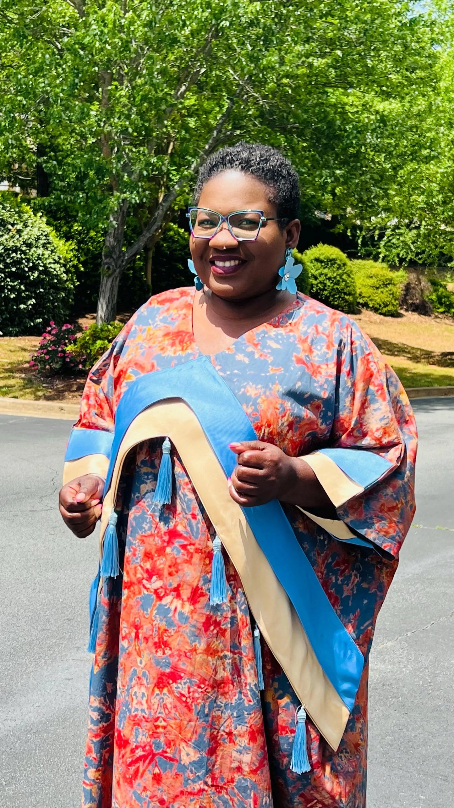 Woman wearing a floor-length Adire and Kampala print dress with angular crepe accents and tassel details, standing outdoors.