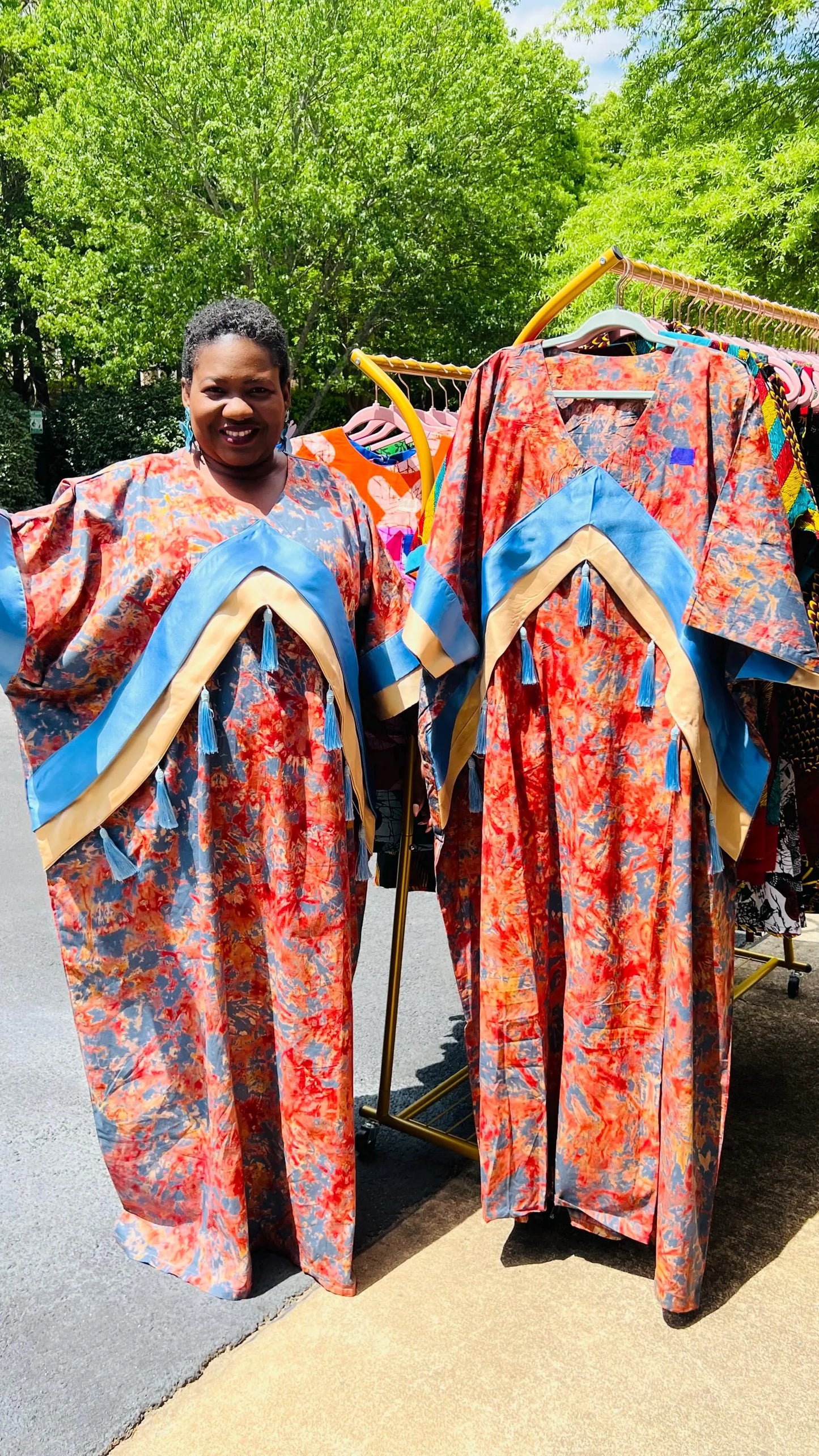 Woman wearing a floor-length Adire and Kampala print dress with angular crepe accents and tassel details, standing outdoors.