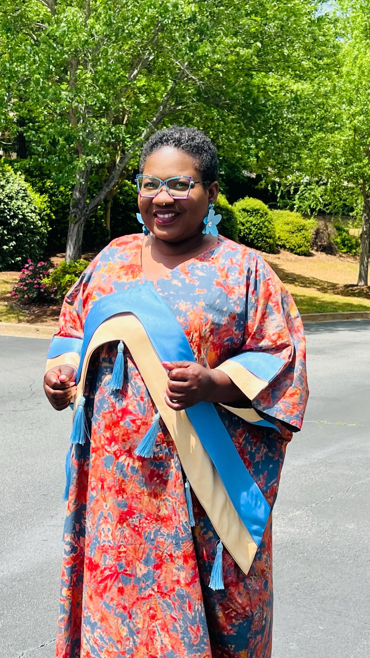 Woman wearing a floor-length Adire and Kampala print dress with angular crepe accents and tassel details, standing outdoors.