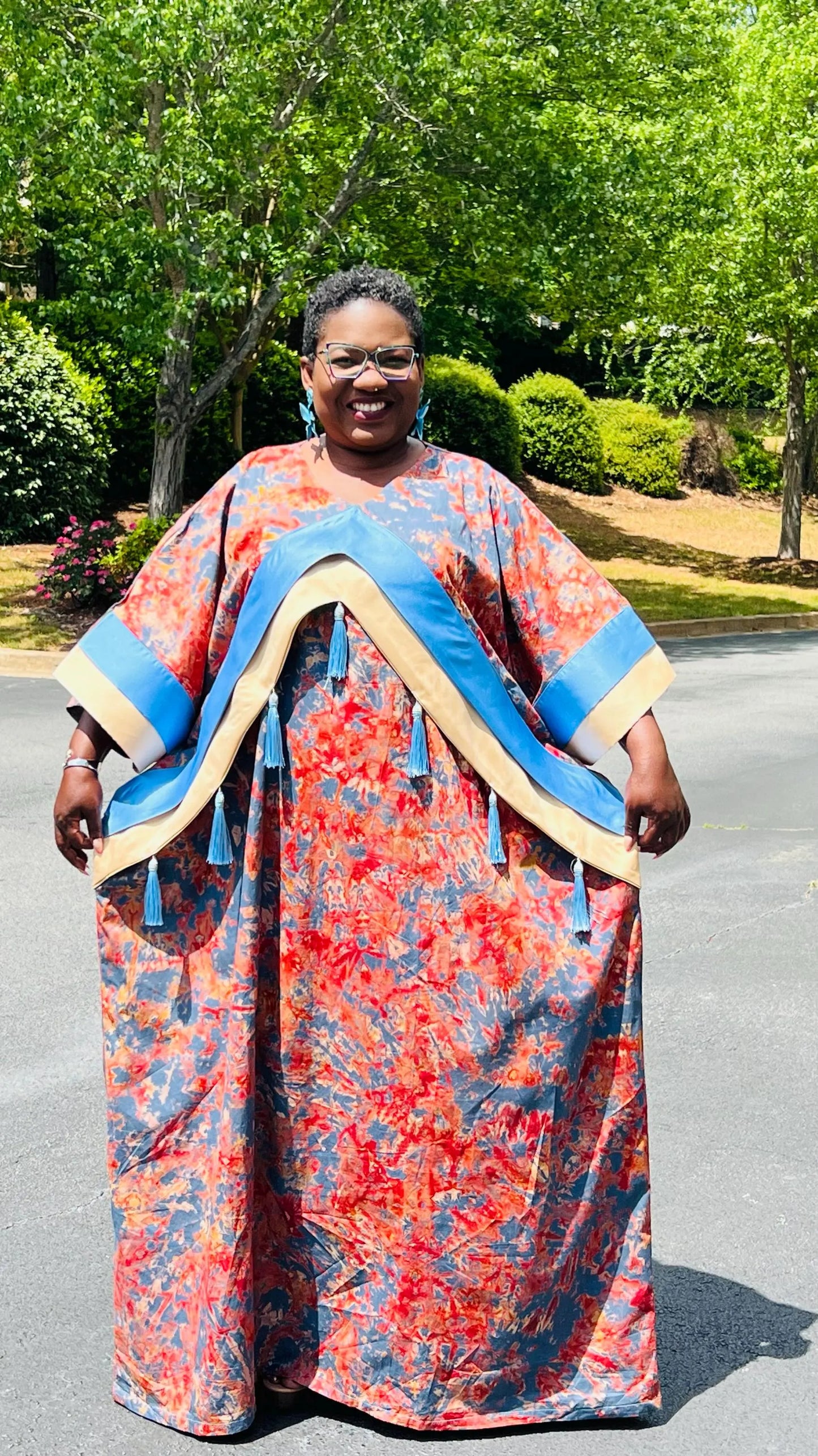 Woman wearing a floor-length Adire and Kampala print dress with angular crepe accents and tassel details, standing outdoors.